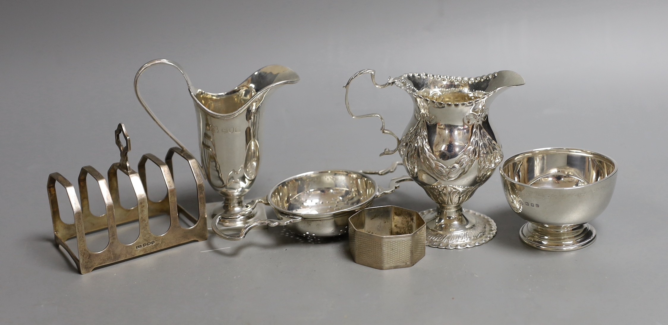 Two late Victorian silver cream jugs, including inverted pear shape, London, 1897, 96mm, a later small silver toastrack, a silver napkin ring and a modern silver tea strainer on stand.